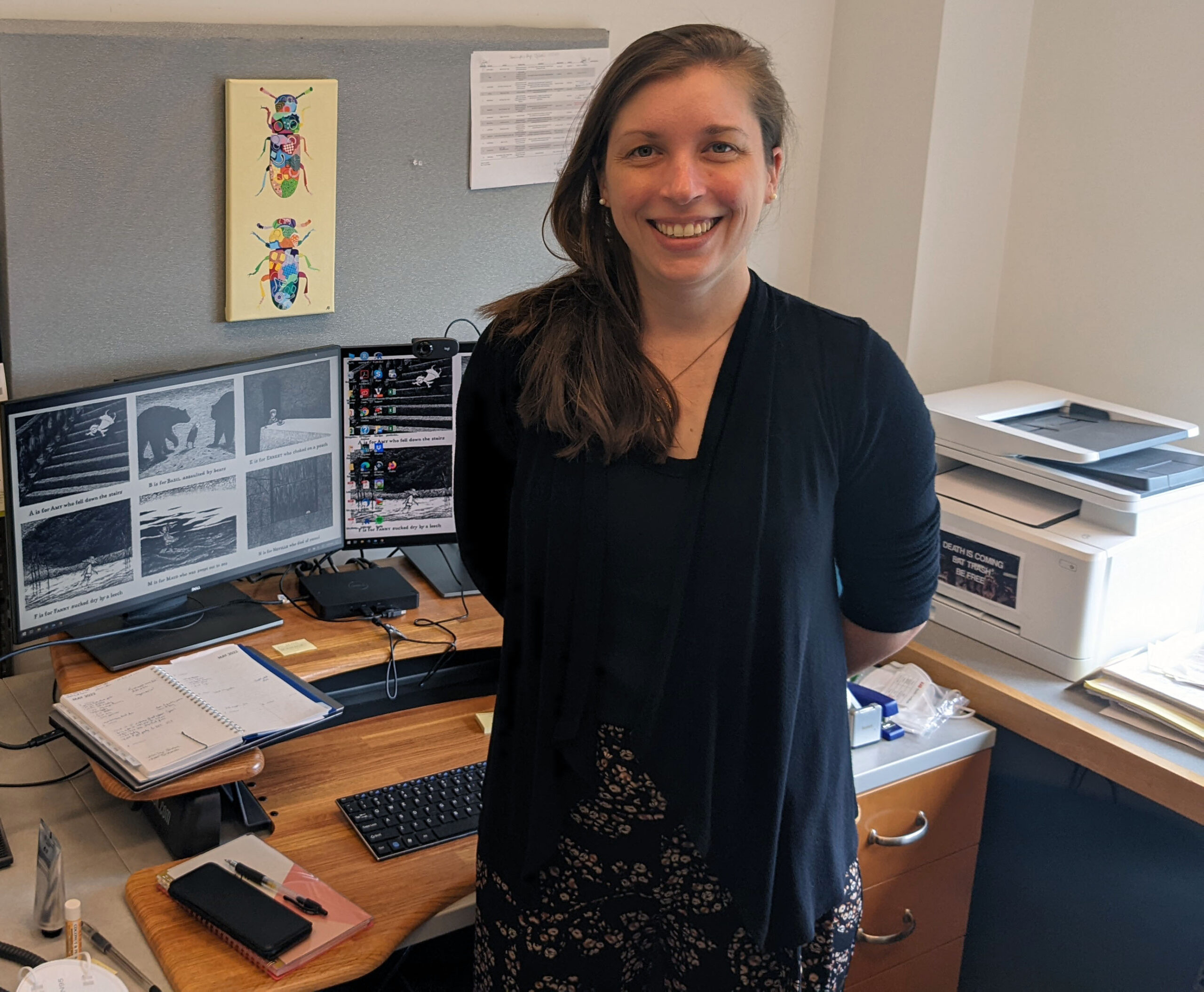 Ann Tate smiling standing in front of her desk with wrap around computer and printer in the background